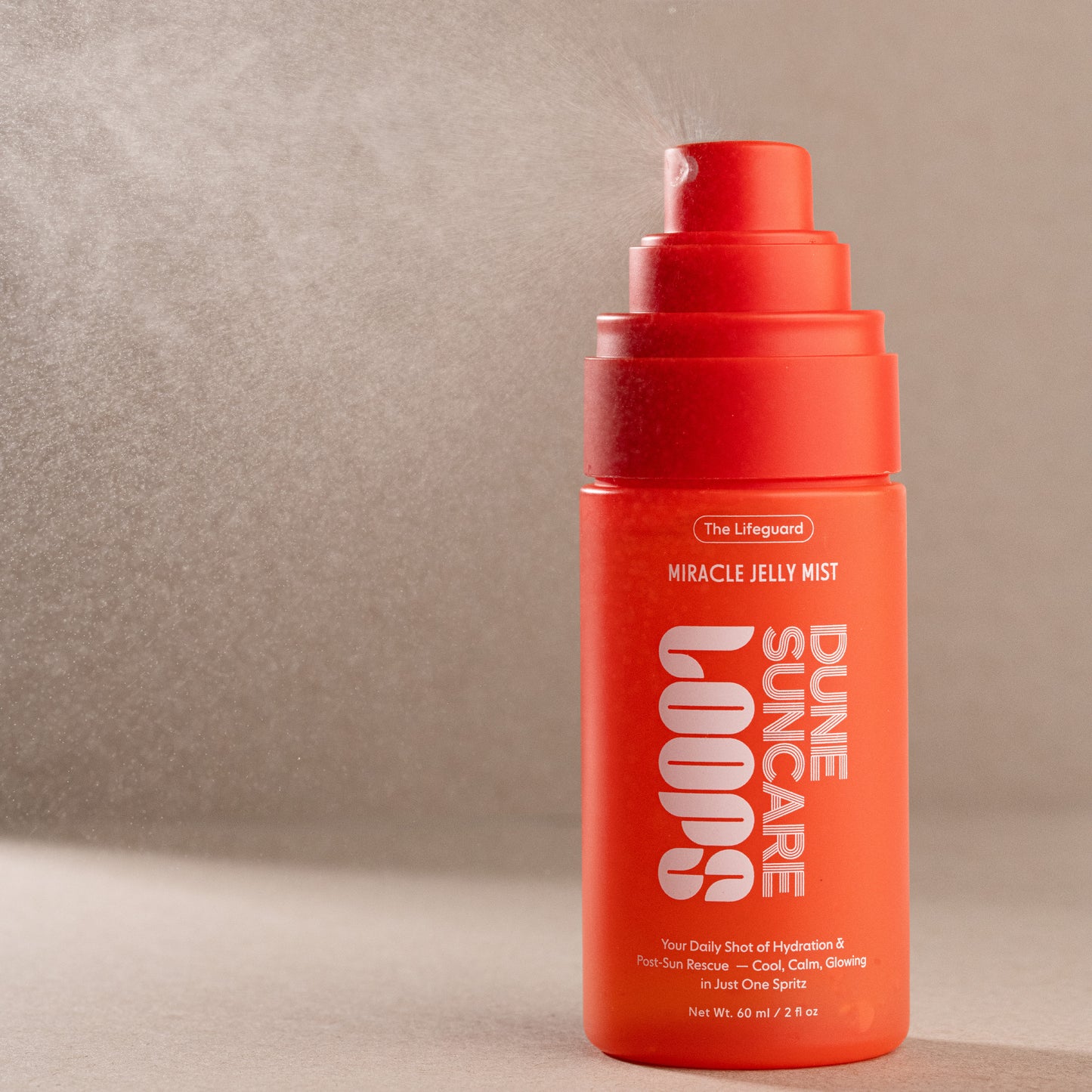 A bright orange-red pump bottle of The Lifeguard Dune Loops Miracle Jelly Mist, shown spraying with a fine mist against a textured beige background.