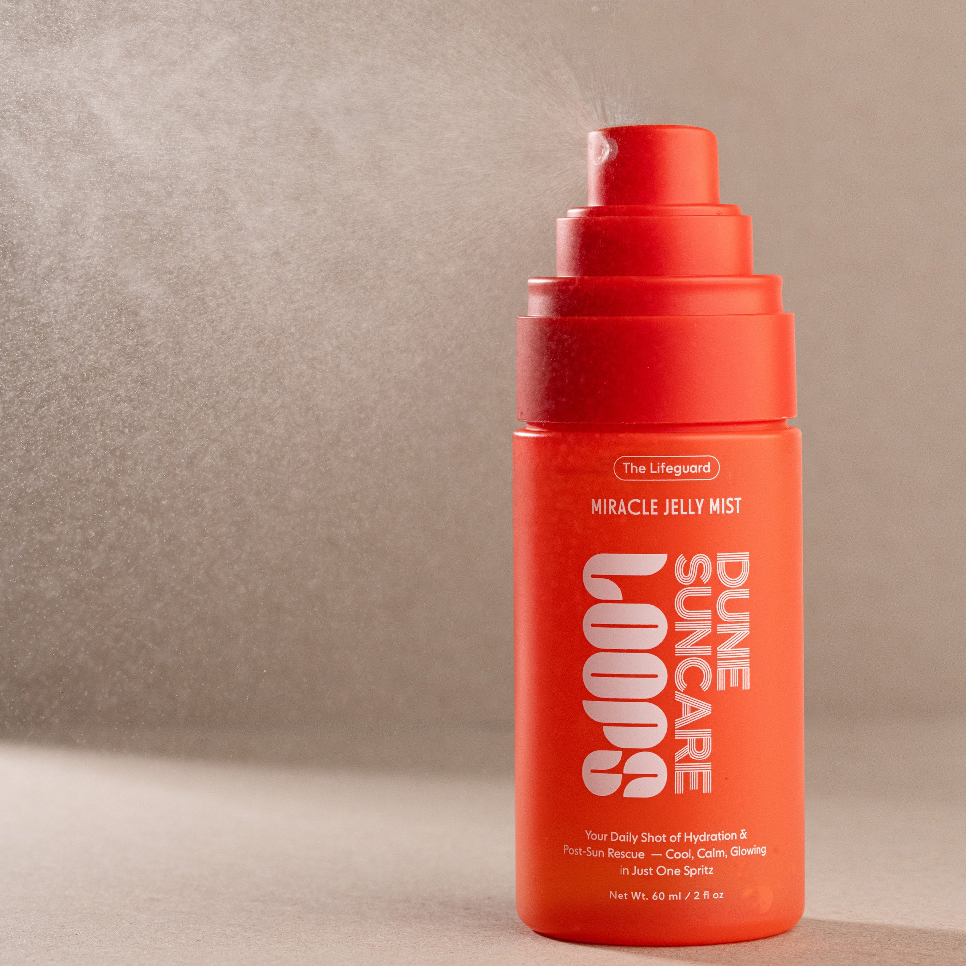 A bright orange-red pump bottle of The Lifeguard Dune Loops Miracle Jelly Mist, shown spraying with a fine mist against a textured beige background.