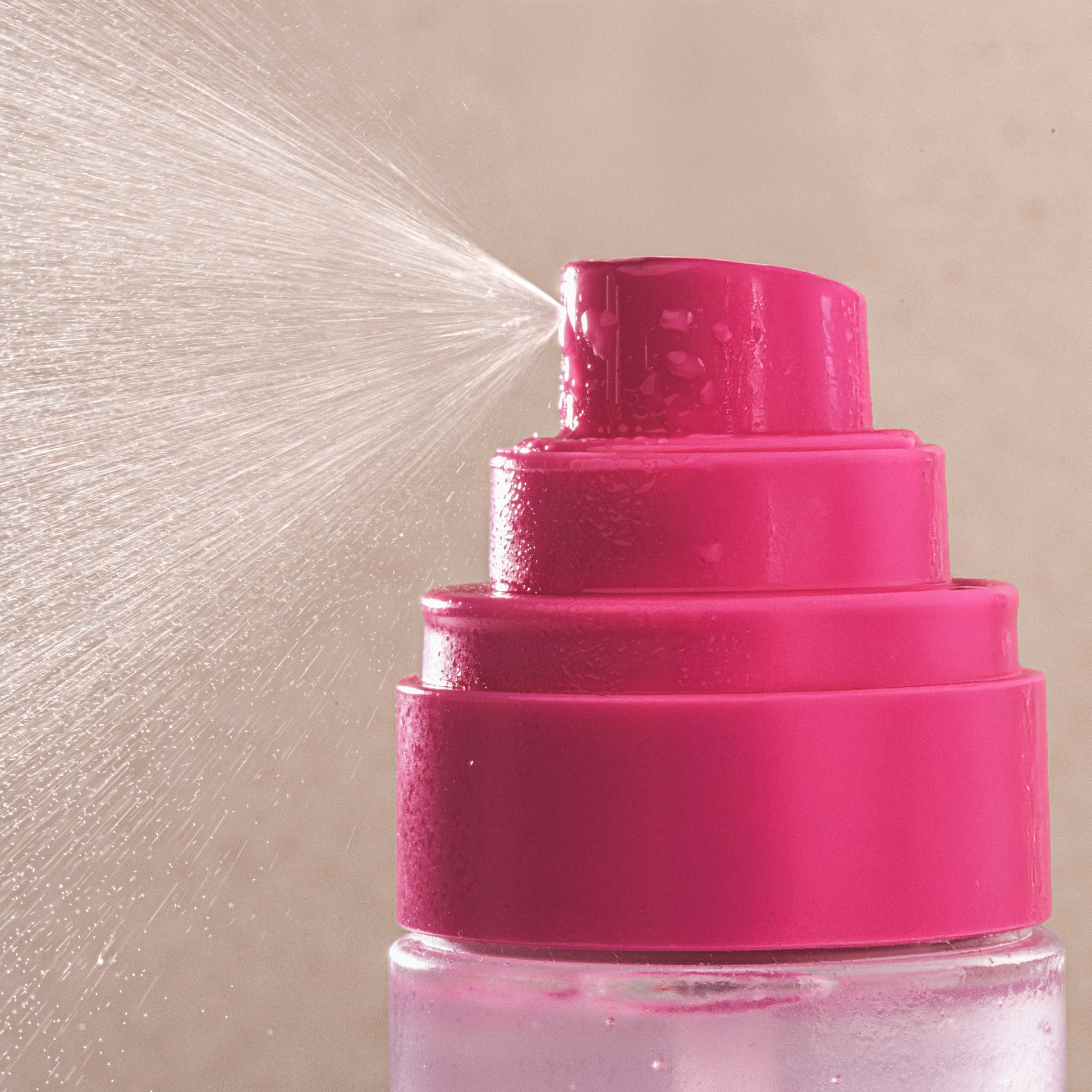 Close-up of a pink spray bottle with water being sprayed against a beige background