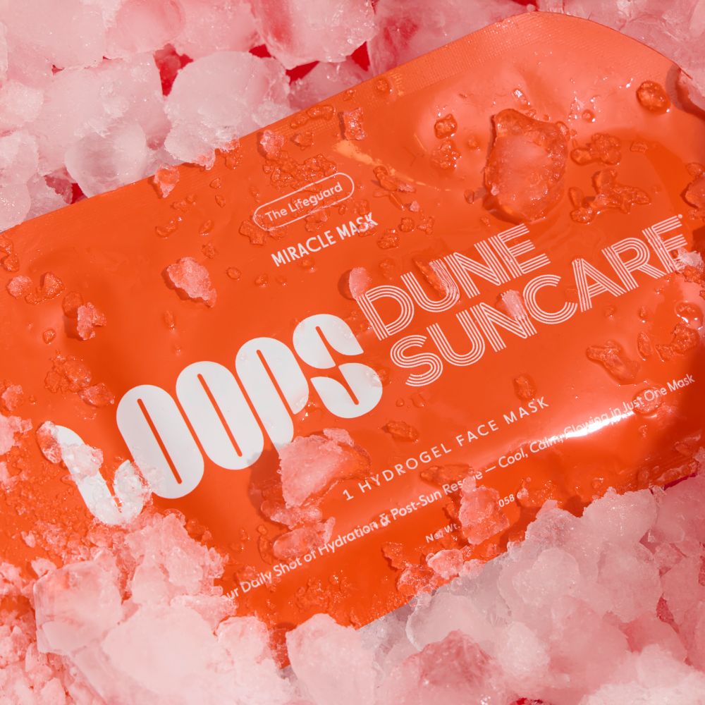 An orange Loops Dune Suncare Miracle Mask face mask packet, covered in water droplets, resting on a bed of pink ice cubes. The brand 'The Lifeguard' is visible on the packaging.