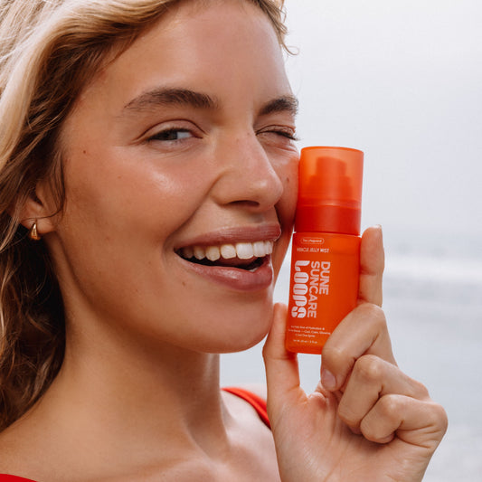 A smiling woman holds an orange bottle of LOOPS x Dune Skincare Miracle Jelly Mist against a soft, bright outdoor background.