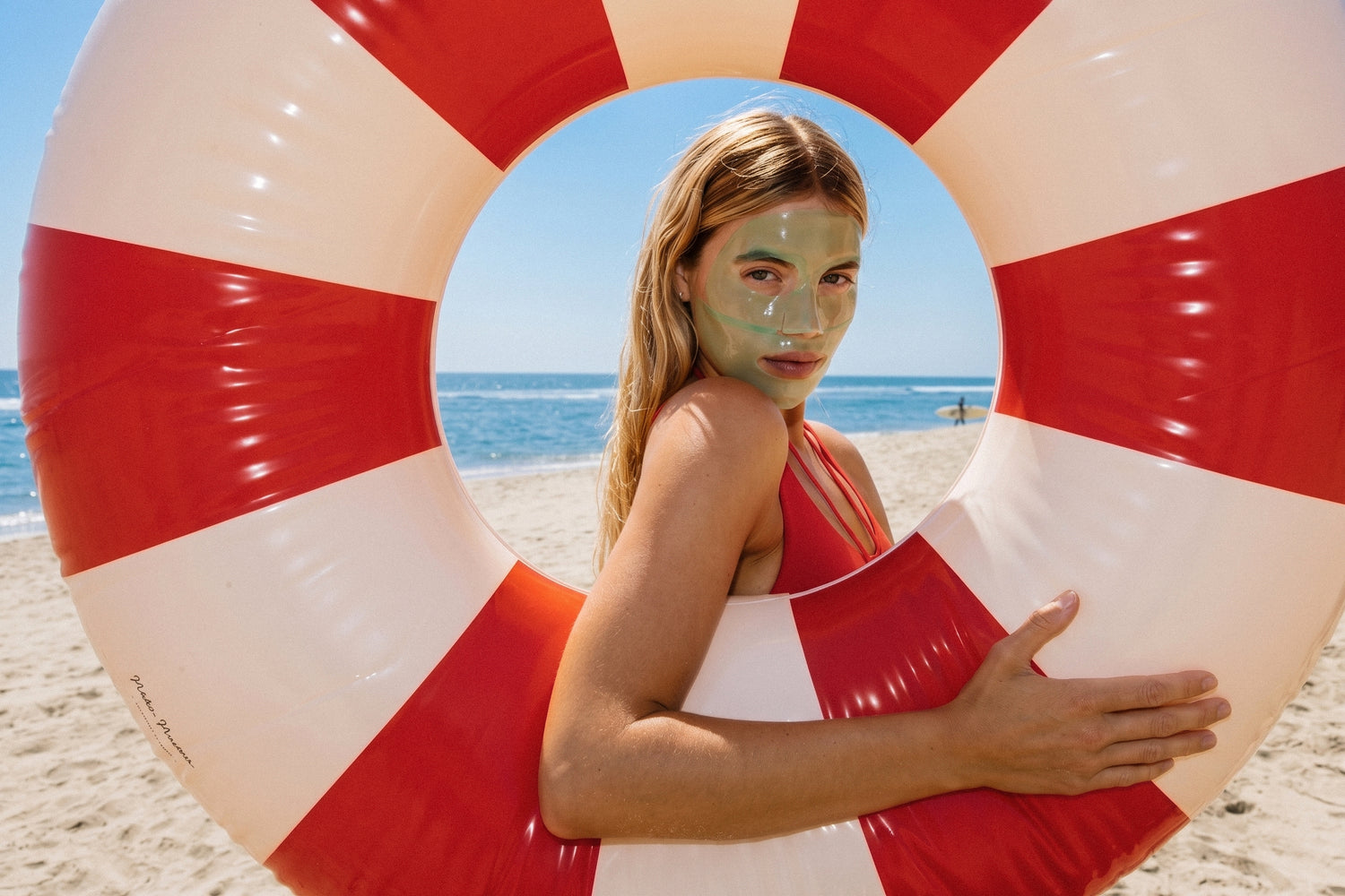 A woman with a green face mask stands on a sandy beach behind a large red and white striped inflatable float