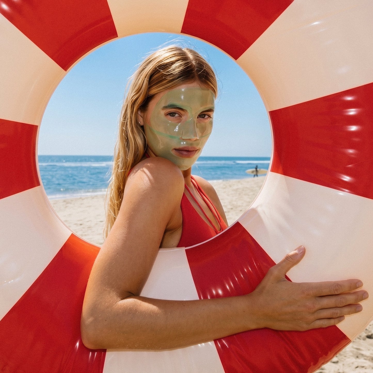 A woman with a green face mask stands on a beach behind a large red and white striped inflatable ring, wearing a red bikini top. The ocean and sand are visible in the background.