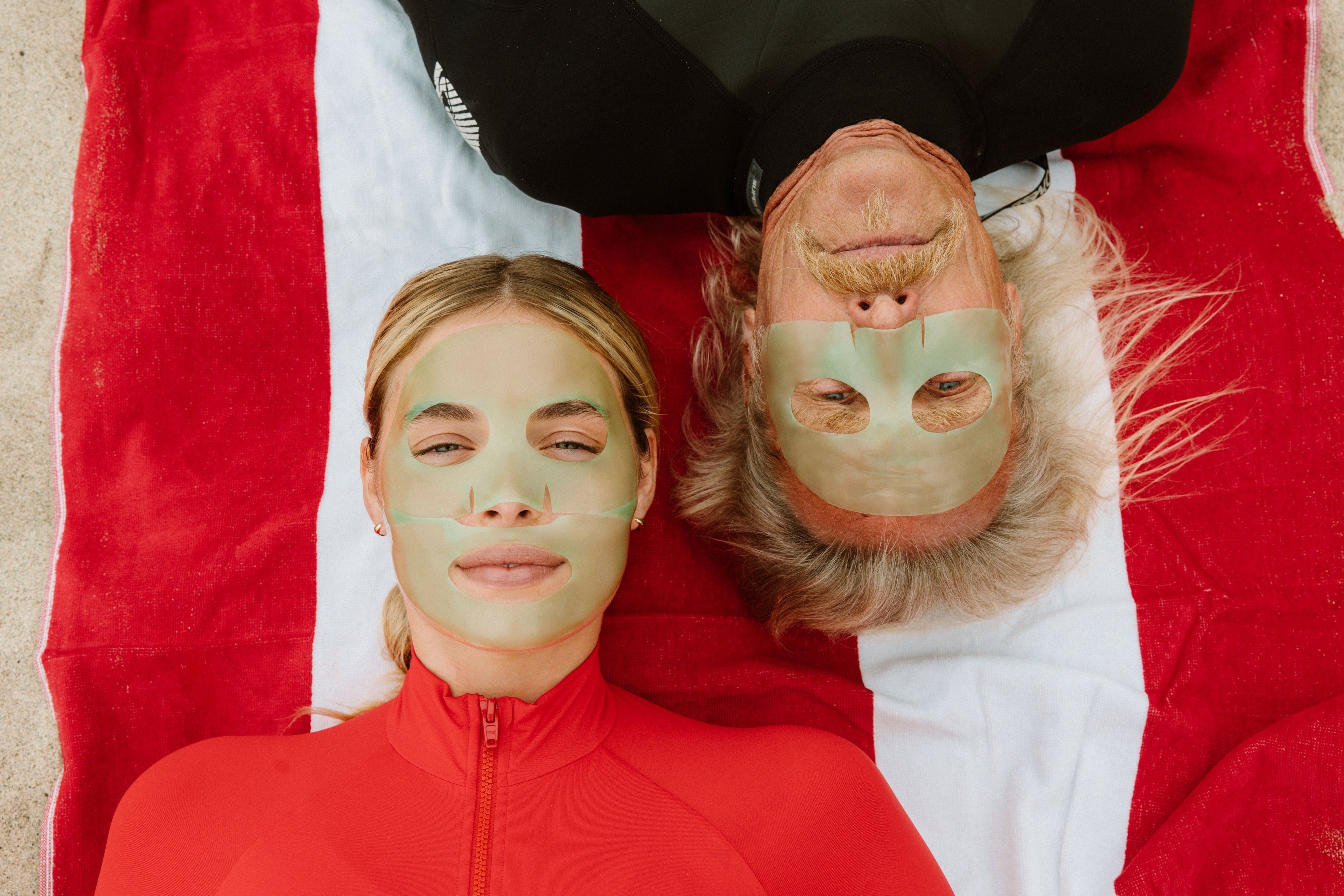 Two women lying side by side on a red and white striped blanket, wearing green face masks and a red zip-up top.